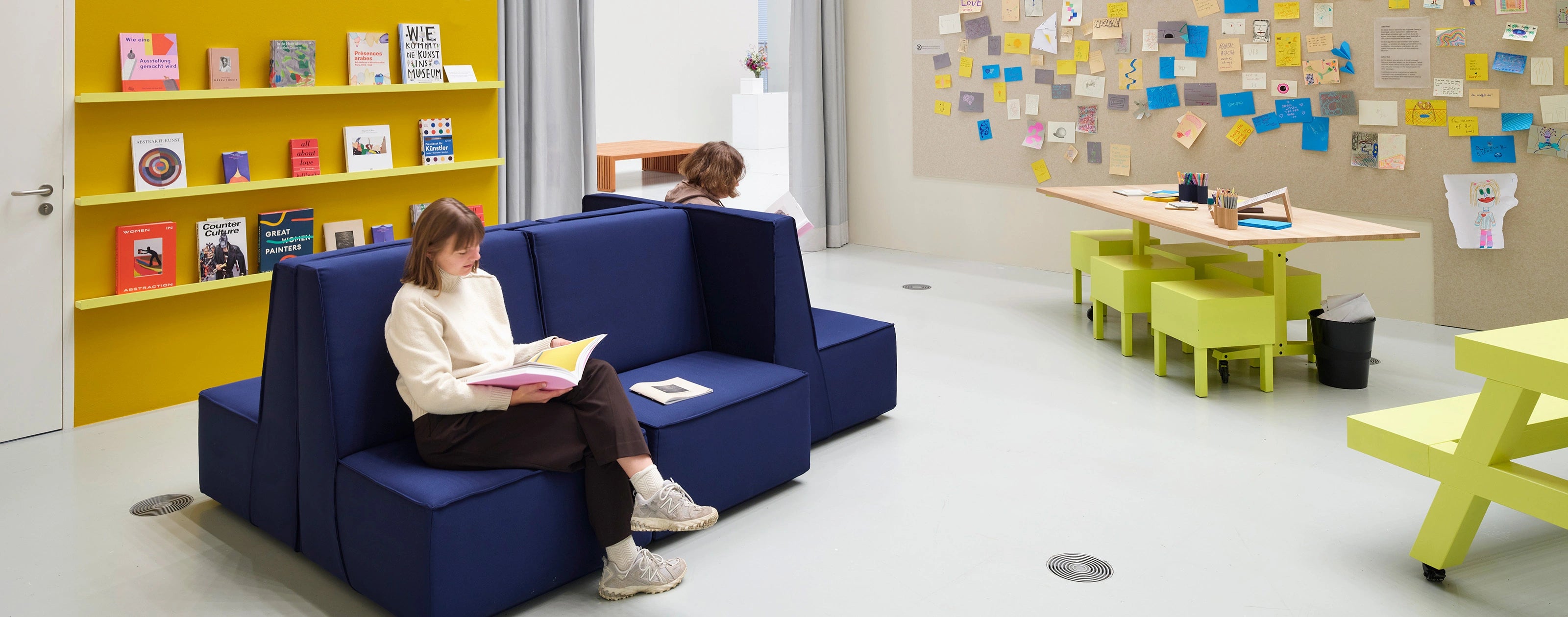 Ein hell gestalteter Raum mit gelben Bücherregalen und einer blauen Sitzbank, auf der eine Frau in einem Buch liest.