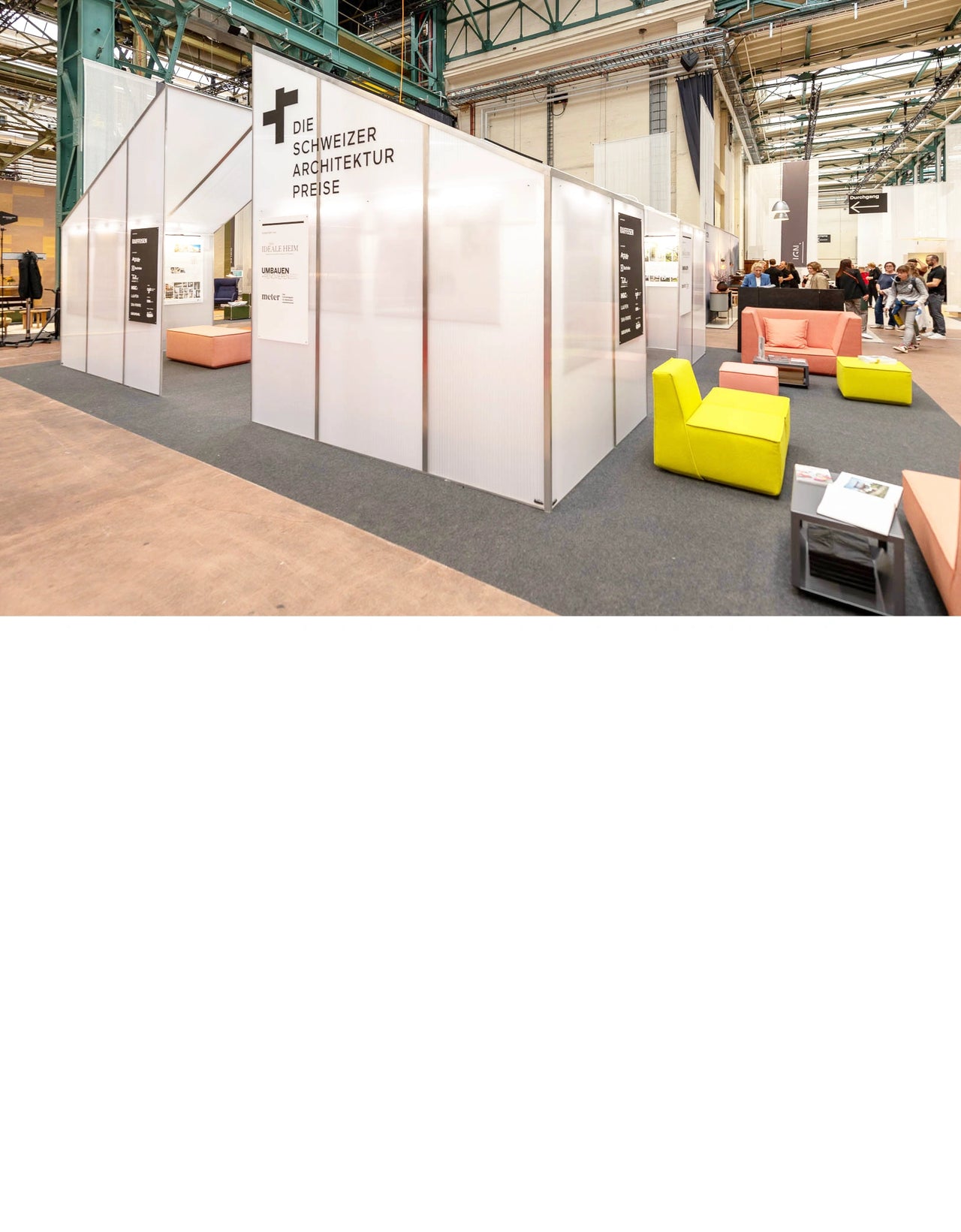 Messestand der Schweizer Architekturpreise in einer Industriehalle, ausgestattet mit modularen Sitzmöbeln in Gelb, Rosa und Orange für eine einladende Lounge-Atmosphäre.