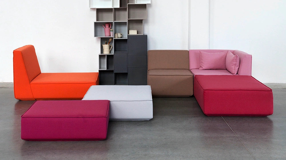 Vielfältige Cubit Sofa-Modul-Kombination in Orange, Pink, Beere und Braun, arrangiert vor einem hohen, grauen Cubit Regalsystem in einem modernen Industrie-Studio.
