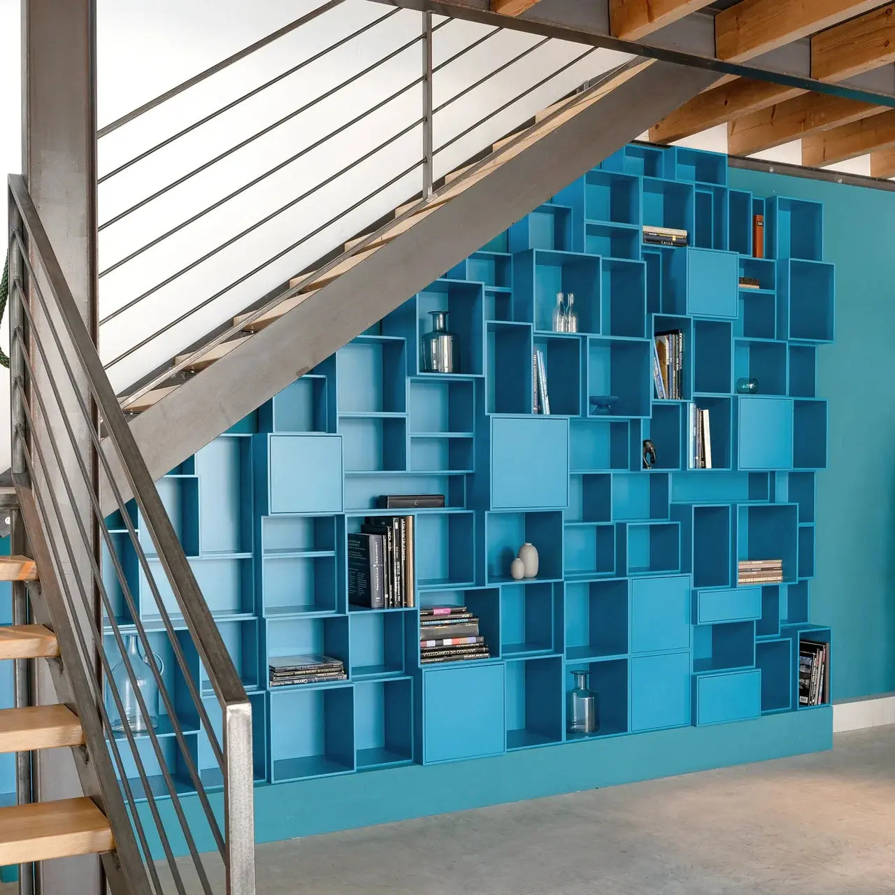 Étagère murale modulaires allant jusqu'au sol, de couleur bleu pétrole, installé sous un escalier ouvert en acier – des cubes personnalisables contenant des livres et des objets de décoration dans un intérieur de loft.