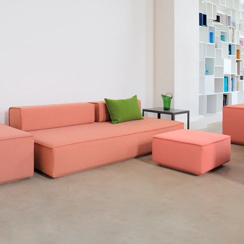 Modulares Schlafsofa in Rosa mit grünem Kissen, Hocker und Beistelltisch vor heller Wand mit weißem Regal im Hintergrund