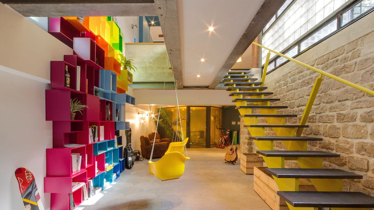 Bright, modern living room with colourful, wall-mounted Shelving made of pink, blue, yellow and green modules, yellow swing and yellow staircase with stone wall and large window front