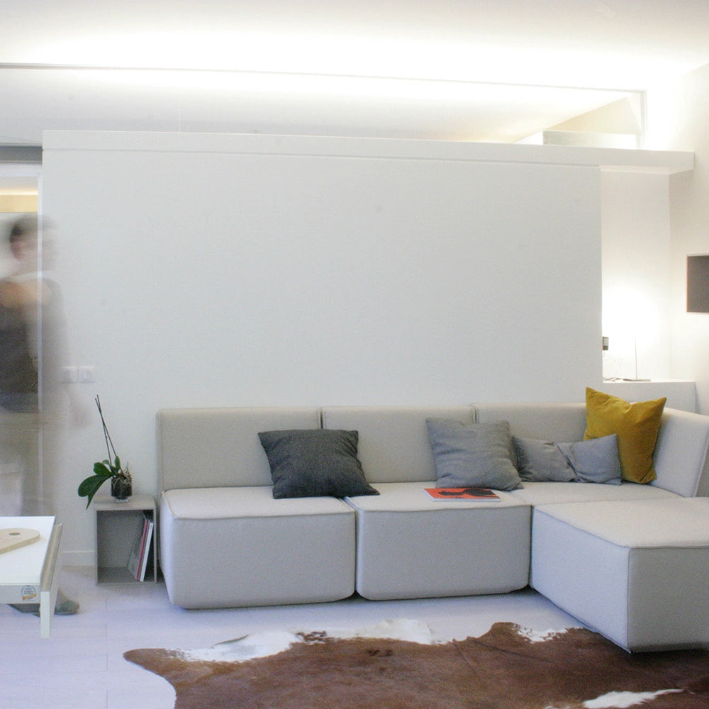 Helles modulares  Ecksofa in Hellgrau mit farbigen Kissen in Anthrazit und Senfgelb vor einer weißen Wand in einem lichtdurchfluteten Raum.