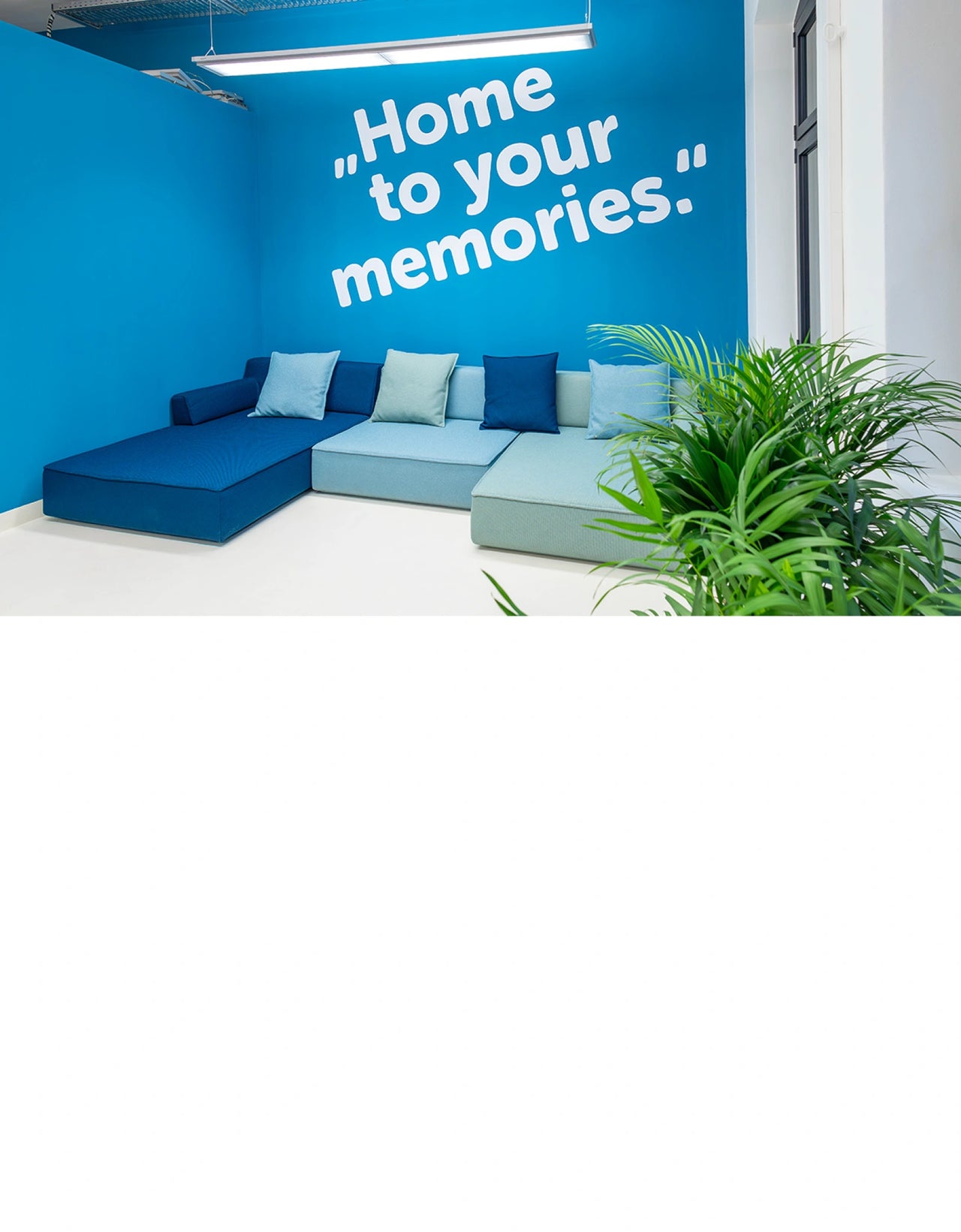 Moderne Lounge mit modularer Sofalandschaft in Blautönen vor einer kräftig blauen Wand mit dem Schriftzug „Home to your memories“, ergänzt durch grüne Zimmerpflanzen im Empfangsbereich.