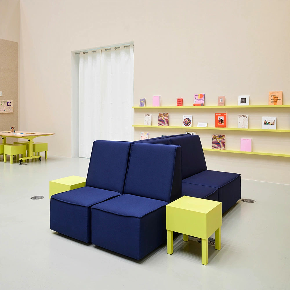 Zweiseitige Cubit Sofa-Insel in Dunkelblau, kombiniert mit neongelben Beistelltischen und Hockern in einem minimalistischen Ausstellungsraum mit gelben Wandregalen.
