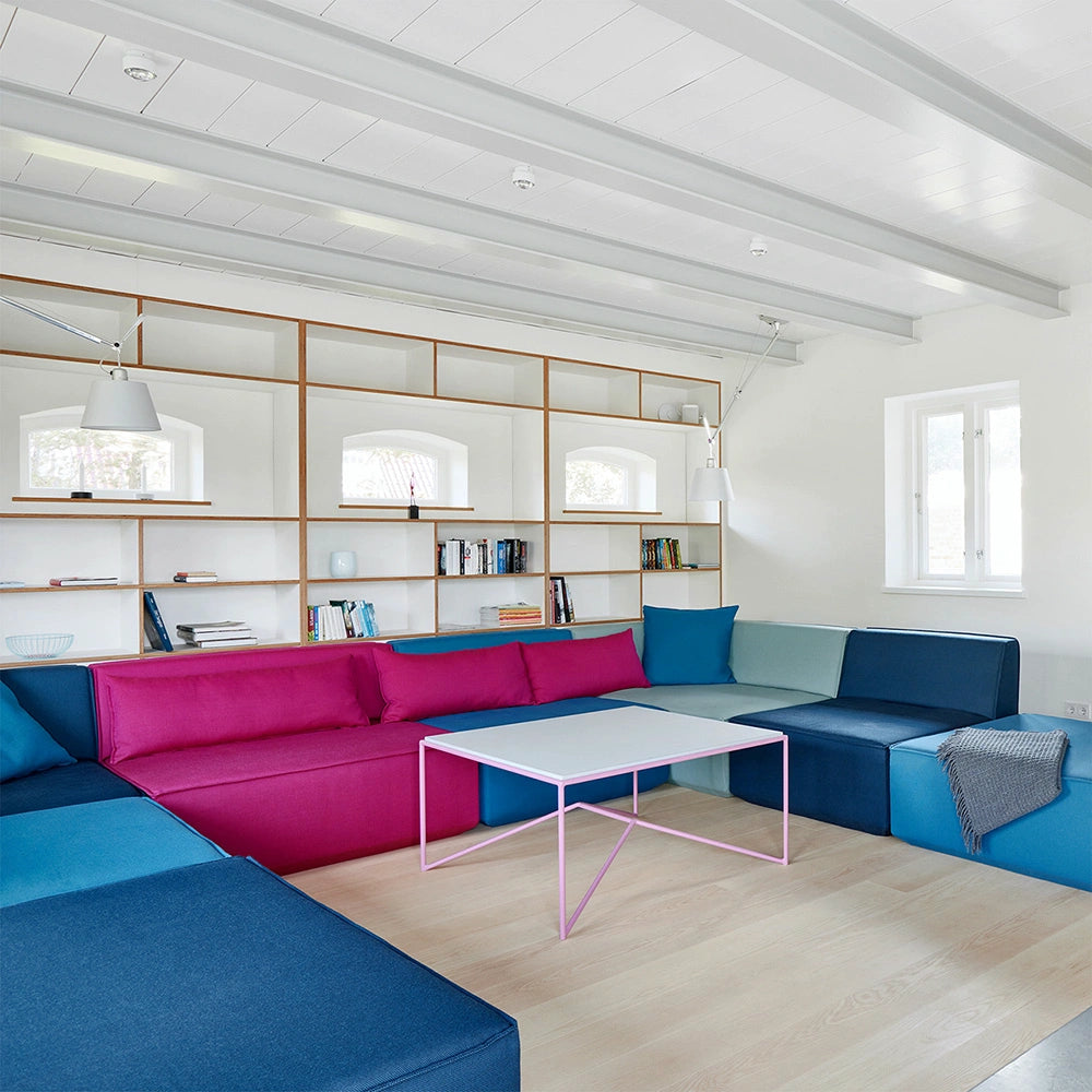 Modernes Wohnzimmer mit einer bunten Cubit Modulsofa-Landschaft in Blau- und Pinktönen vor einem minimalistischen weißen Wandregal.